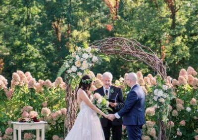Elegant outdoor wedding ceremony in lush green garden with floral arch and bride and groom exchanging vows.